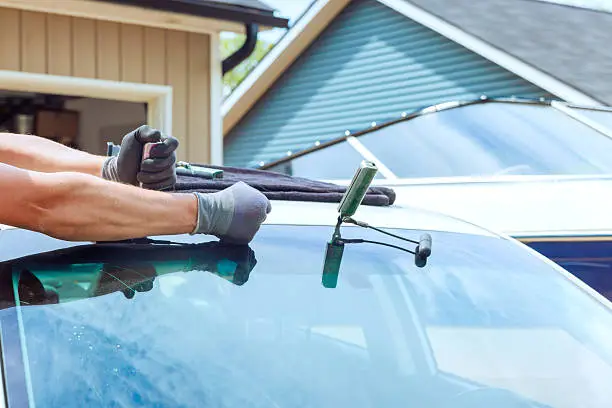 Person in gloves applying sealant to BMW windshield edge with car in foreground.