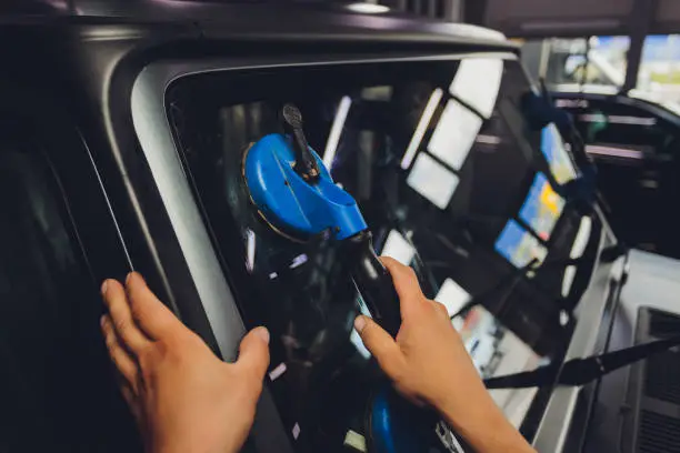 A person using a blue suction cup tool to handle a car windshield, with hands pressing against the glass.