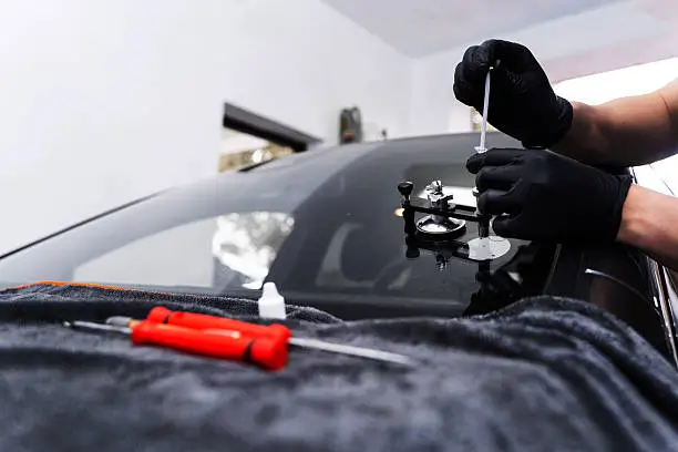 A person wearing black gloves uses a syringe and suction tool to repair a car windshield, with tools on a cloth nearby.
