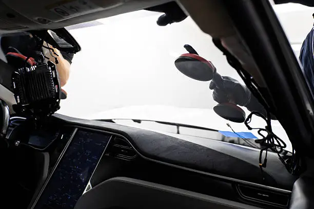 Inside a car, a person uses red-handled suction cups to work on the windshield, with a dashboard and equipment visible.