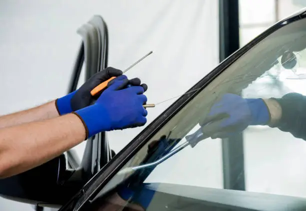 Technician wearing blue gloves uses a tool to remove or install the windshield of a car inside a workshop, working along the car’s roofline.